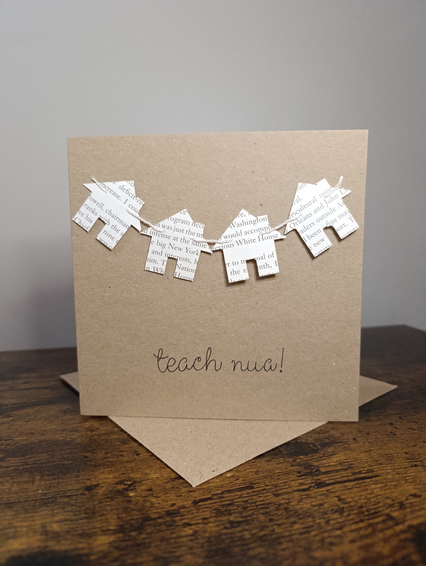 Kraft paper card with paper houses and 'teach nua!' text on a wooden surface.
