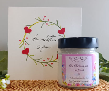 Jar of soy wax candle from Sliabh candles with a decorative card featuring hearts and text on a textured surface. An Mháthair is fearr which means the best mam. 