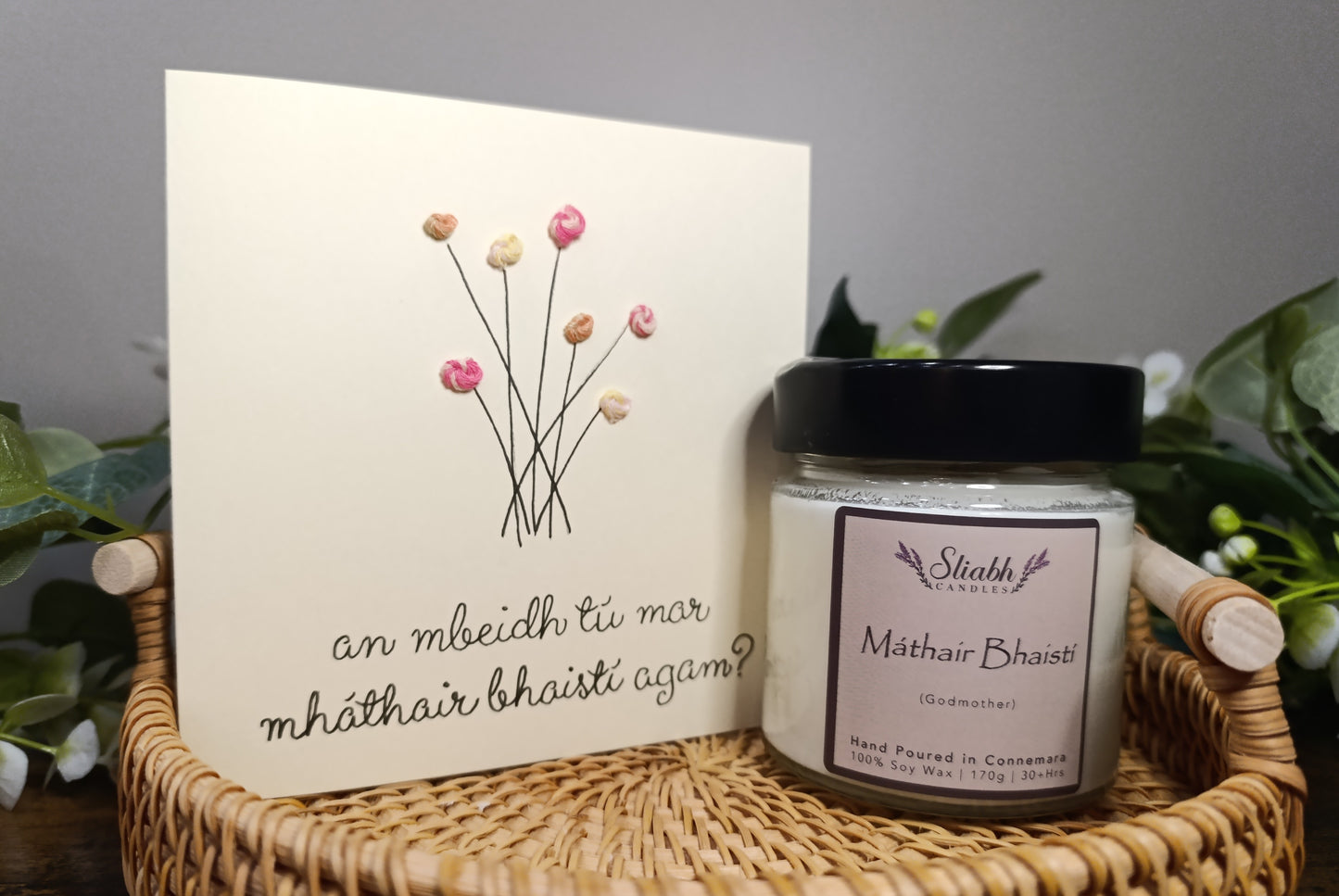 Irish Candle with Máthair Bhaistí and  Irish card on a woven basket with plants in the background.  Godmother proposal gift set. 