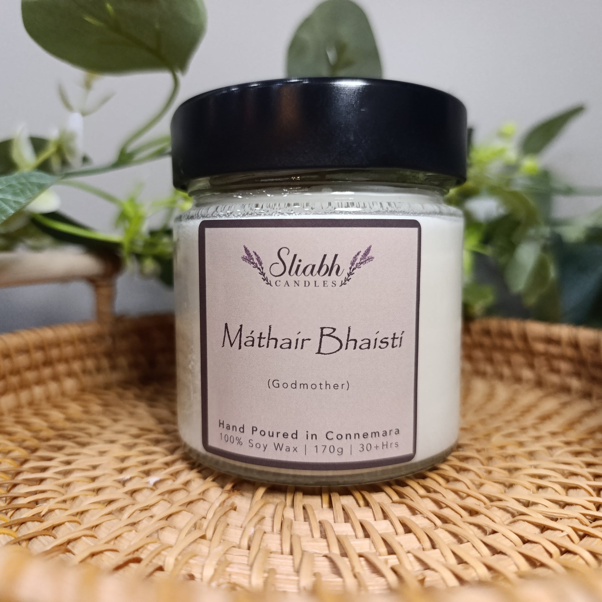 Jar of 'Mathair Bhaisti' candle on a woven tray with plants in the background. Godmother Gift Set