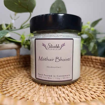 Jar of 'Mathair Bhaisti' candle on a woven tray with plants in the background. Godmother Gift Set