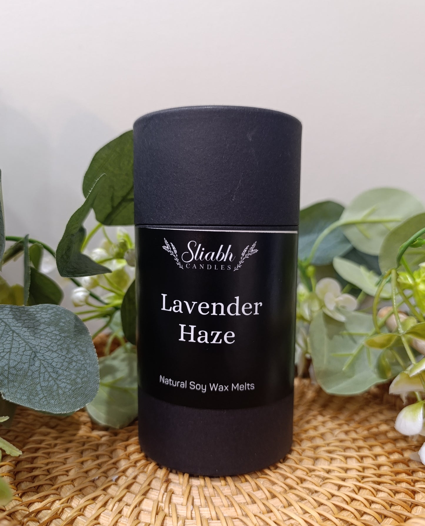 Black container of 'Lavender Haze' soy wax melts with 'Sliabh Candles' branding on a woven surface with greenery. Irish wax melts handmade in Connemara. Lavender scented.