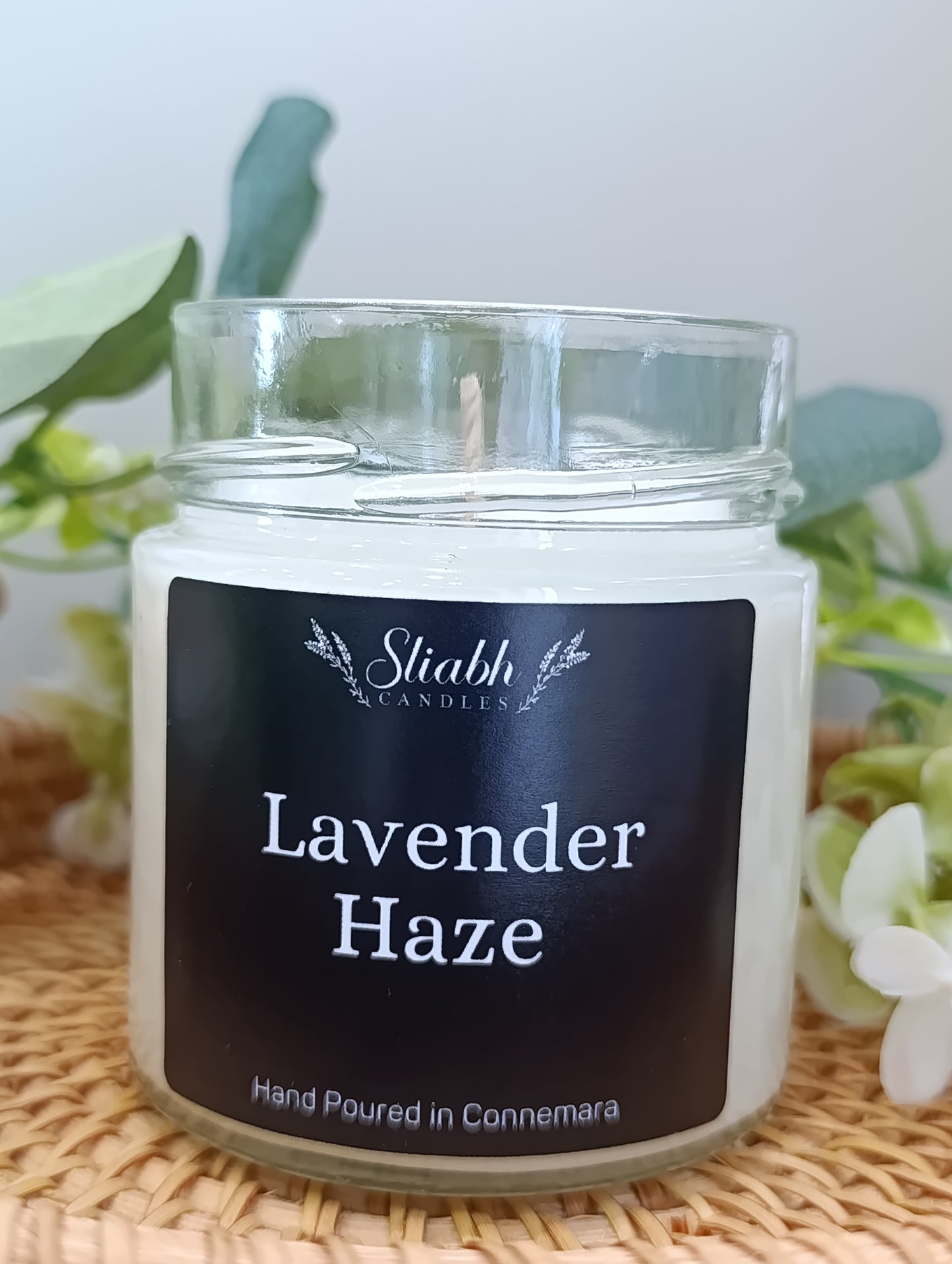 Candle jar labeled 'Lavender Haze' by Sliabh Candles on a woven surface with greenery in the background.