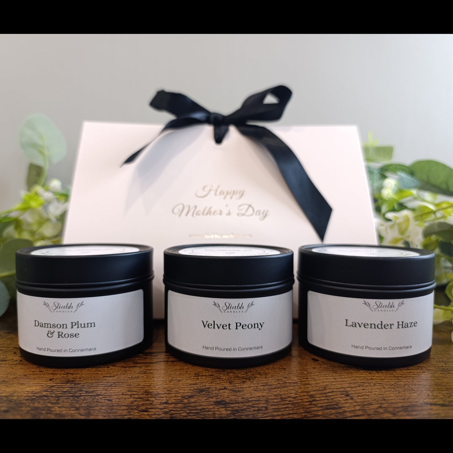 Three small jars with black lids and white labels on a wooden surface, with a gift box and flowers in the background.