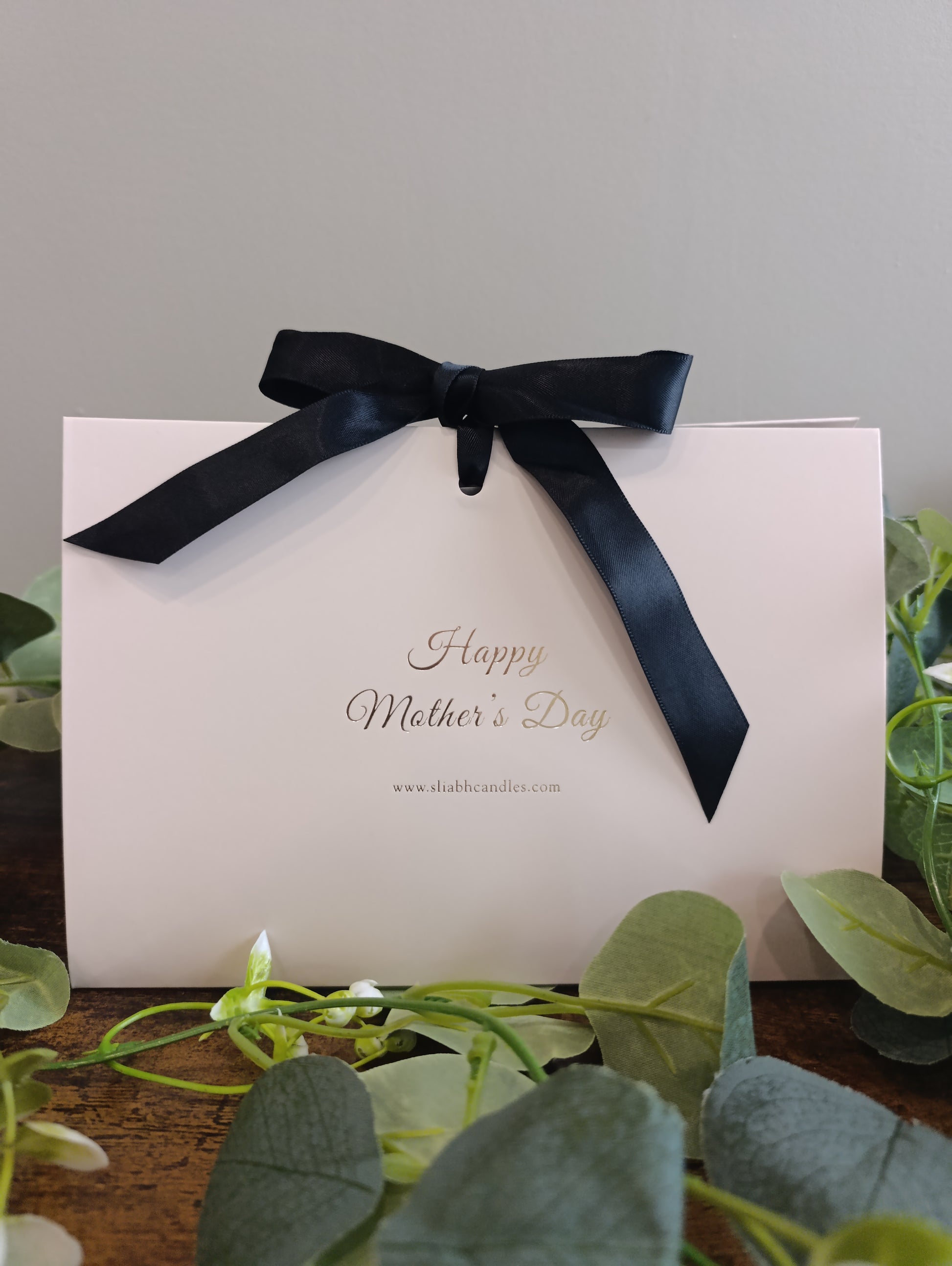 White box with a black ribbon and 'Happy Mother's Day' text, surrounded by greenery.
