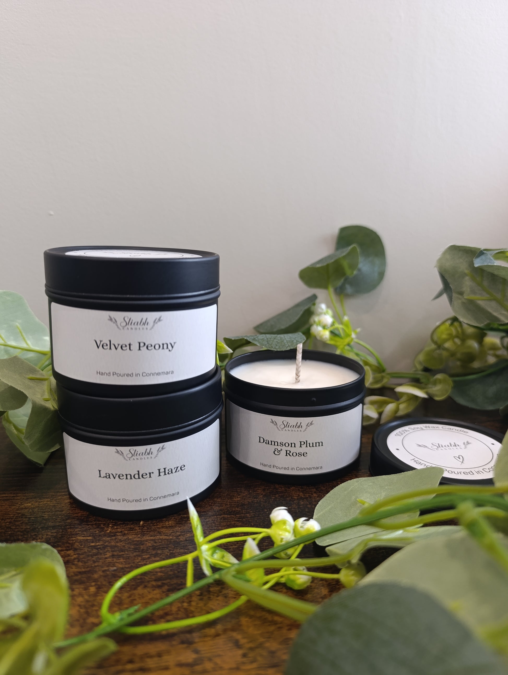 Three candles with labels on a wooden surface surrounded by green leaves