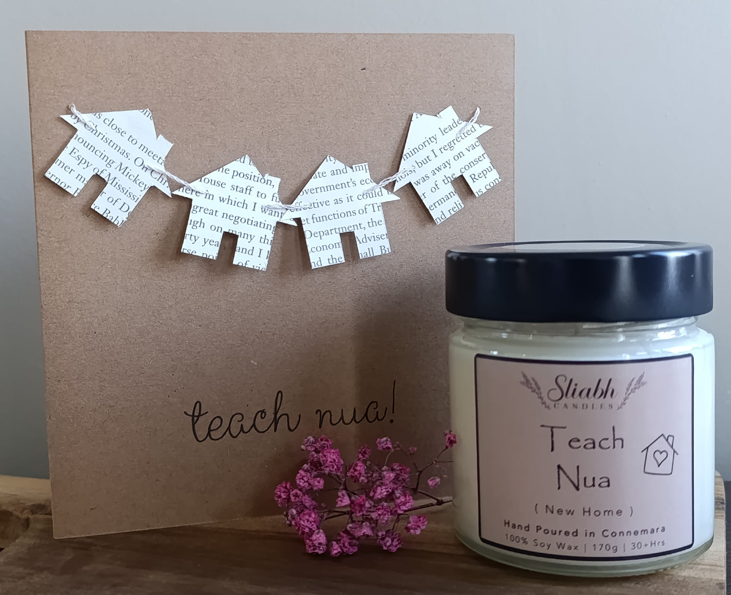 Candle labeled 'Teach Nua' with decorative paper houses on a brown card.