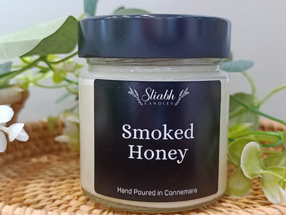 Jar of smoked honey candle with a black lid on a woven surface with plants in the background