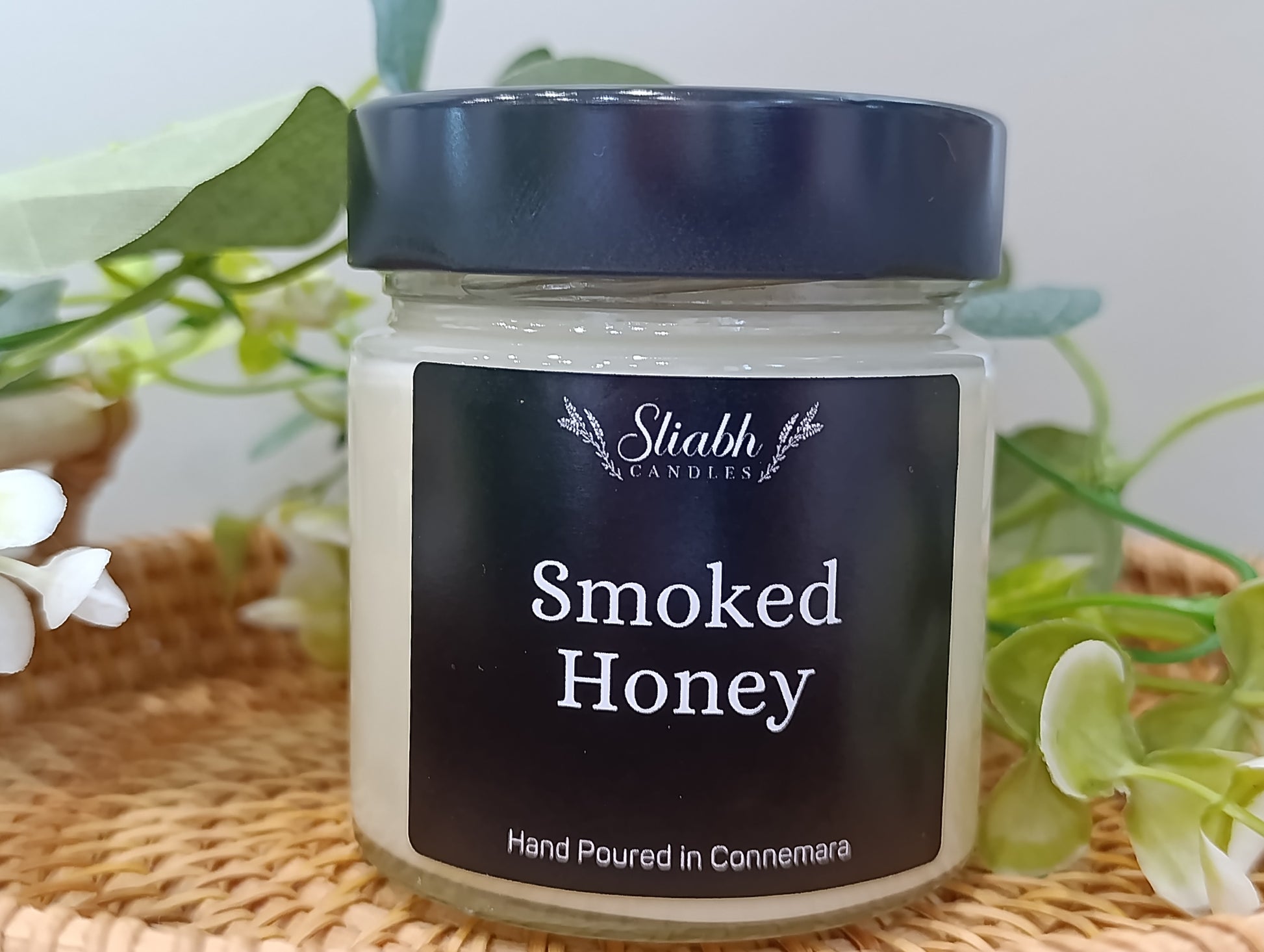 Jar of smoked honey with a black lid on a woven surface, surrounded by greenery.