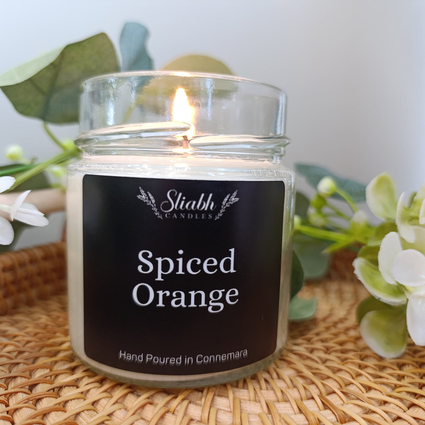 Candle labeled 'Spiced Orange' by Slabh Candles on a woven surface with flowers in the background.