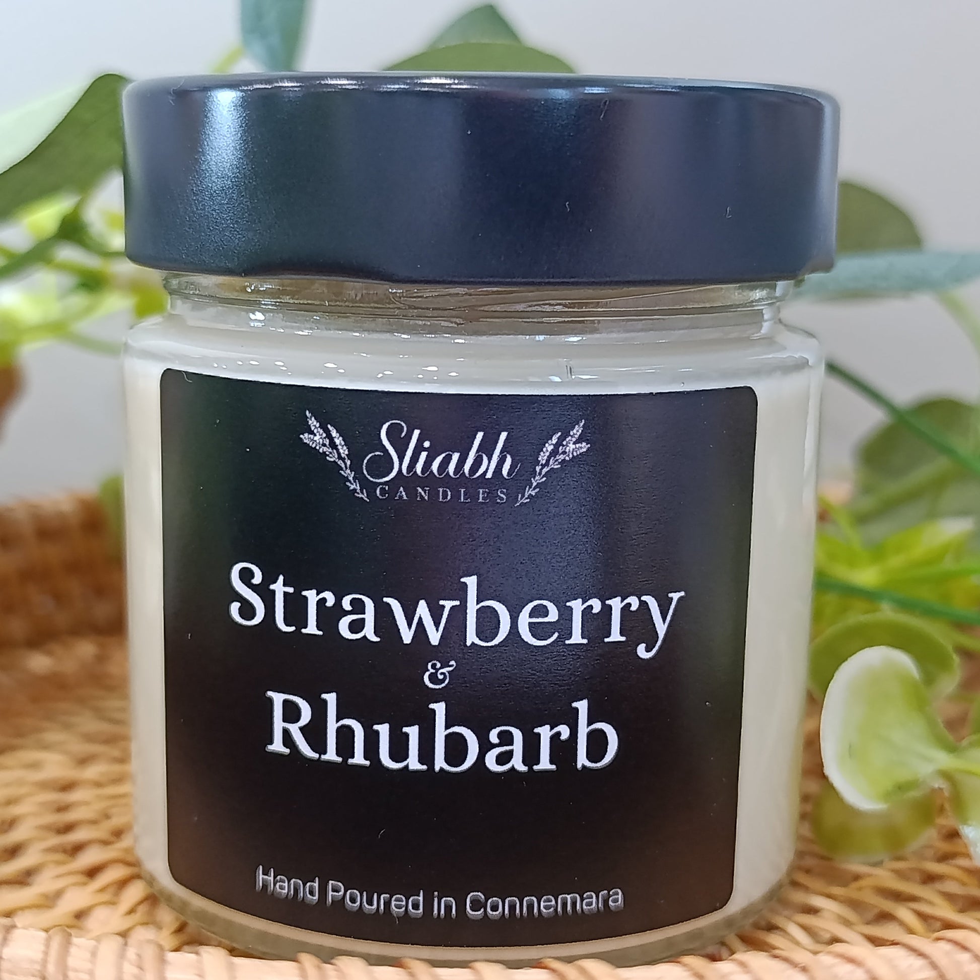 Jar of 'Strawberry & Rhubarb' candle with a black lid and label on a woven surface.