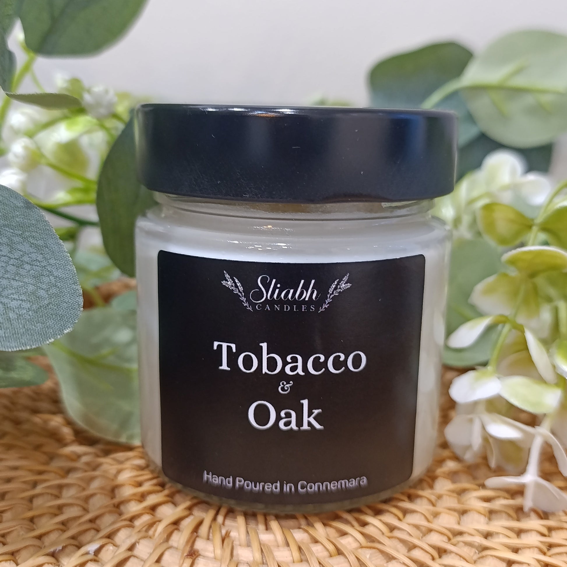 Jar of 'Tobacco & Oak' candle with a black lid on a woven surface with greenery.