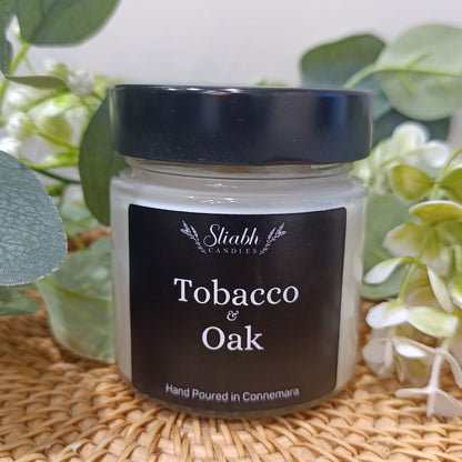 Jar of 'Tobacco & Oak' candle with a black lid on a woven surface with greenery.