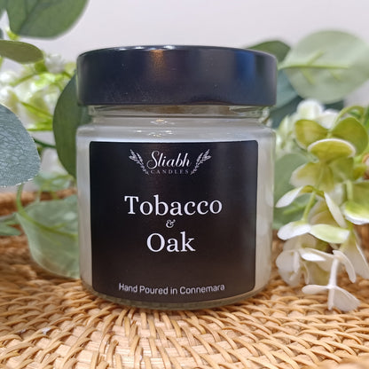 Candle jar labeled 'Tobacco & Oak' by Sliabh Aromatics on a woven surface with greenery.