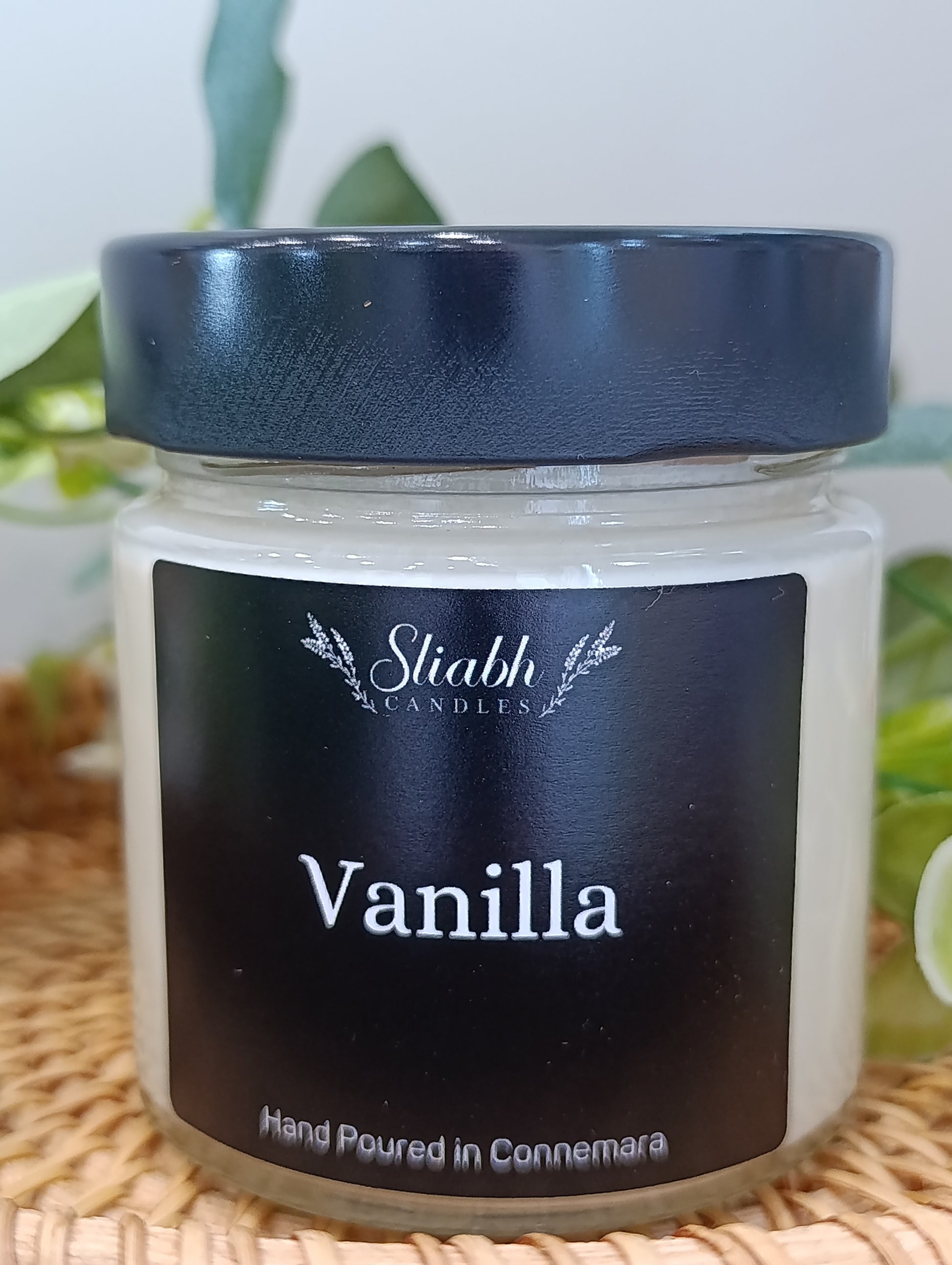 Vanilla scented candle jar with black lid and label on a woven surface.